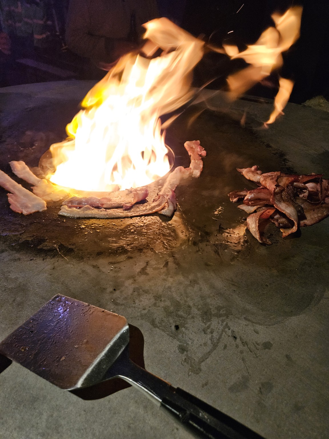 Speck wird über offenem Feuer auf der Feuerplatte gebraten
